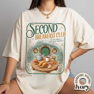 May include: Ivory-colored t-shirt with a green and brown graphic. The graphic features the text "SECOND BREAKFAST CLUB" above an illustration of a hobbit hole with a table set for breakfast. The shirt also has the words "for the looking to share in an adventure".