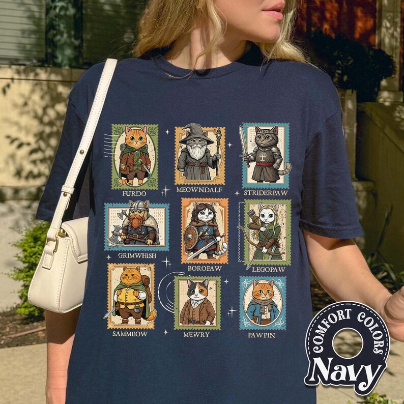 K&ouml;nnte beinhalten: Marineblaues T-Shirt mit neun Katzenillustrationen in Briefmarkendesigns. Jede Katze ist in Fantasy-Kleidung gekleidet, mit Namen wie "Furdo" und "Meowndalf". Auf dem Shirt stehen die Worte "Comfort Colors" und "Navy".