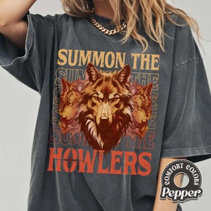 May include: Dark grey t-shirt featuring a wolf graphic with the text "SUMMON THE HOWLERS" in orange and tan. The design includes three wolf heads. The shirt has a vintage, worn look.