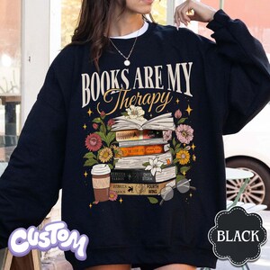 May include: Black sweatshirt with the text "BOOKS ARE MY Therapy" above a stack of books and flowers. The books have titles like "Rebecca Yarros" and "Fourth Wing." A coffee cup and glasses are also featured.