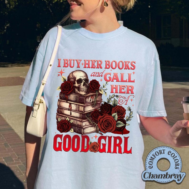 May include: Light blue t-shirt with a graphic featuring a skull atop a stack of books, surrounded by red roses. The text reads "I BUY HER BOOKS and GALL HER GOOD GIRL". The shirt is from Comfort Colors.