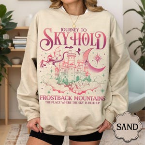 May include: Sand-colored sweatshirt with a fantasy castle graphic. The design features a castle, dragon, and crescent moon with the text "Journey to Sky Hold" and "Frostback Mountains". The sweatshirt is a crew neck style.
