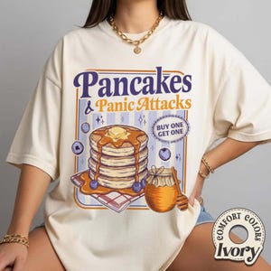 May include: Ivory Comfort Colors t-shirt with a graphic of pancakes, syrup, blueberries, and a honey pot. Text reads "Pancakes & Panic Attacks" with a "Buy One Get One" offer.
