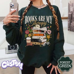 May include: Forest green sweatshirt with the text "BOOKS ARE MY Therapy" above a stack of books, flowers, and a coffee cup. The books have titles like "Rebecca Yarros" and "Fourth Wing". The sweatshirt has a custom design.