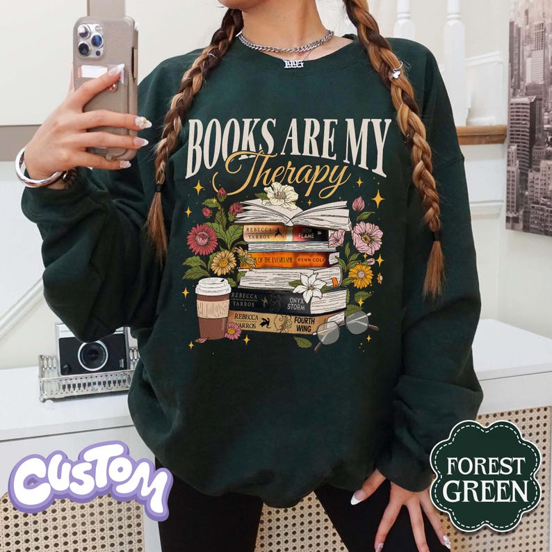 May include: Forest green sweatshirt with the text "BOOKS ARE MY Therapy" above a stack of books, flowers, and a coffee cup. The books have titles like "Rebecca Yarros" and "Fourth Wing". The sweatshirt has a custom design.
