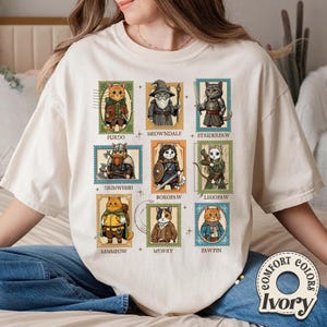 K&ouml;nnte beinhalten: Elfenbeinfarbenes Comfort Colors T-Shirt mit neun Katzenillustrationen im Stil von Briefmarken. Jede Katze ist in Fantasy-Kleidung gekleidet, mit Namen wie Furdo, Meowndalf und Striderpaw. Das Shirt hat eine lockere Passform und ein Comfort Colors-Label.