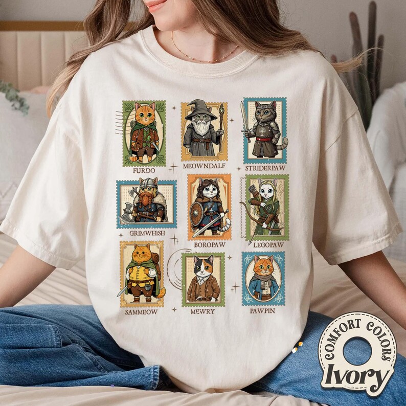 K&ouml;nnte beinhalten: Elfenbeinfarbenes Comfort Colors T-Shirt mit neun Katzenillustrationen im Stil von Briefmarken. Jede Katze ist in Fantasy-Kleidung gekleidet, mit Namen wie Furdo, Meowndalf und Striderpaw. Das Shirt hat eine lockere Passform und ein Comfort Colors-Label.