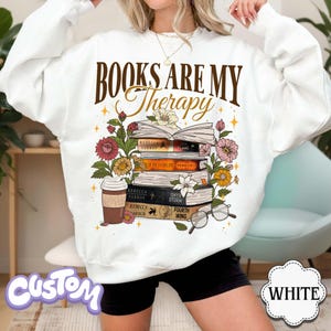 May include: White sweatshirt with the text "BOOKS ARE MY Therapy" in brown script. The design includes a stack of books, flowers, a coffee cup, and glasses. The sweatshirt is a comfortable, casual style.