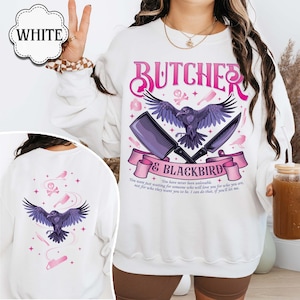 May include: White sweatshirt with a graphic design featuring a raven, cleaver, and knife, with the text "BUTCHER & BLACKBIRD". The back of the sweatshirt also has a raven graphic and text. The design includes pink and purple accents.
