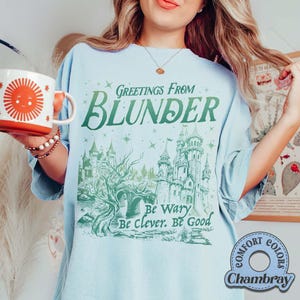 Puede incluir: Camiseta azul claro con el texto "Greetings From Blunder" y una ilustraci&oacute;n de castillo verde. La camiseta tambi&eacute;n presenta las frases "Be Wary, Be Clever, Be Good." En la imagen se sostiene una taza roja y blanca con un dise&ntilde;o de sol.