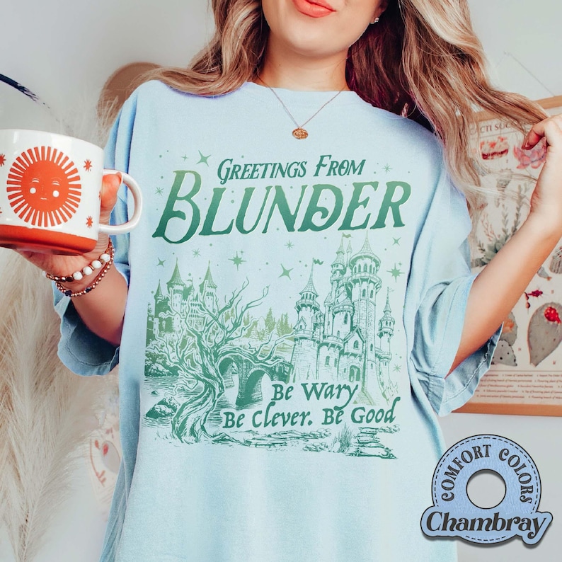 Puede incluir: Camiseta azul claro con el texto "Greetings From Blunder" y una ilustraci&oacute;n de castillo verde. La camiseta tambi&eacute;n presenta las frases "Be Wary, Be Clever, Be Good." En la imagen se sostiene una taza roja y blanca con un dise&ntilde;o de sol.