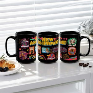 May include: Three black ceramic mugs with colorful graphic designs. Each mug features unique text and illustrations, including phrases like "You Will Not Break Me" and "New Achievement!" The mugs have large handles and are displayed on a white surface.