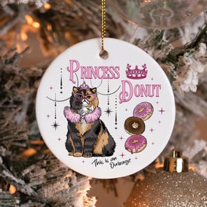 May include: A white, round Christmas ornament with a cat illustration wearing glasses, a crown, and a pink boa. The ornament reads "Princess Donut" with images of donuts and the phrase "This is an Outrage".