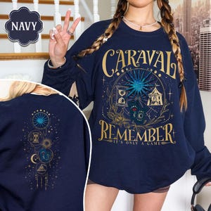 K&ouml;nnte beinhalten: Marineblaues Sweatshirt mit goldenem und t&uuml;rkisfarbenem Grafikdesign. Die Vorderseite zeigt den Text "CARAVAL REMEMBER" mit einer himmlischen und zirkusartigen Illustration. Die R&uuml;ckseite hat ein kleineres passendes Design.