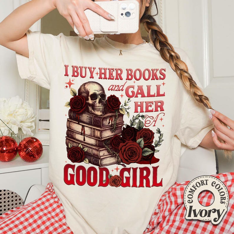 May include: Cream-colored t-shirt with a graphic design featuring a skull atop a stack of books, surrounded by red roses. The text reads "I BUY HER BOOKS and GALL HER GOOD GIRL". The shirt has a Comfort Colors Ivory label.