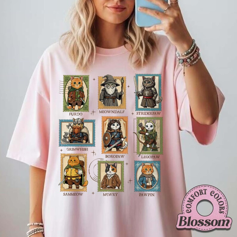 K&ouml;nnte beinhalten: Hellrosa T-Shirt mit neun Katzenillustrationen in Briefmarkendesigns. Jede Katze ist in Fantasy-Kleidung gekleidet, mit Namen wie "Furdo" und "Meowndalf". Das Shirt hat ein "Comfort Colors"-Logo unten rechts.