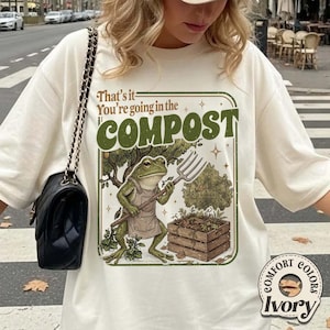 May include: Ivory-colored t-shirt with a graphic of a frog holding a garden fork, wearing an apron, and standing near a compost bin. The text on the shirt reads "That's it - You're going in the COMPOST."