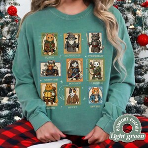 K&ouml;nnte beinhalten: Hellgr&uuml;nes Sweatshirt mit neun Katzenillustrationen in Briefmarkendesigns. Jede Katze ist in Fantasy-Kleidung gekleidet und tr&auml;gt Namen wie Furdo, Meowndale und Striderpaw. In der unteren rechten Ecke befindet sich ein rundes Logo mit der Aufschrift "Comfort Colors" und "Light green".