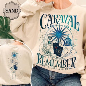 K&ouml;nnte beinhalten: Sandfarbenes Sweatshirt mit teal- und blauem Grafikdesign. Das Design enth&auml;lt das Wort "CARAVAL" &uuml;ber "REMEMBER", mit einem Riesenrad, einer Sanduhr, Spielkarten und einem Zelt. Die R&uuml;ckseite des Sweatshirts hat ein passendes Design.
