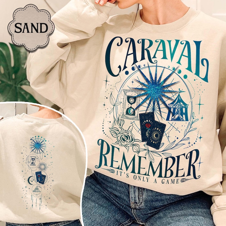 K&ouml;nnte beinhalten: Sandfarbenes Sweatshirt mit teal- und blauem Grafikdesign. Das Design enth&auml;lt das Wort "CARAVAL" &uuml;ber "REMEMBER", mit einem Riesenrad, einer Sanduhr, Spielkarten und einem Zelt. Die R&uuml;ckseite des Sweatshirts hat ein passendes Design.