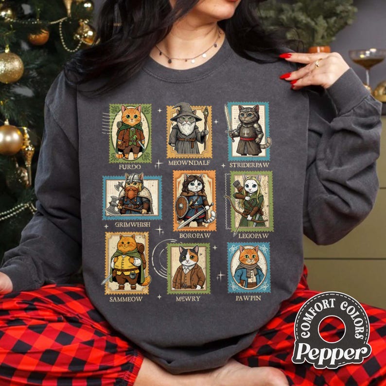 K&ouml;nnte beinhalten: Dunkelgraues Sweatshirt mit neun Katzenillustrationen in Briefmarkendesigns. Jede Katze ist in Fantasy-Kleidung gekleidet und tr&auml;gt Namen wie "Furdo" und "Meowndale". Das Sweatshirt hat die Logos "Comfort Colors" und "Pepper".