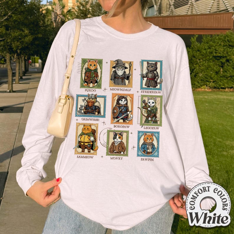 K&ouml;nnte beinhalten: Wei&szlig;es Langarmshirt mit neun Katzenillustrationen, die als Briefmarken gestaltet sind. Jede Katze ist in einem einzigartigen Kost&uuml;m dargestellt, mit Namen wie "Furdo" und "Meowndalf". Das Shirt hat ein "Comfort Colors"-Logo.