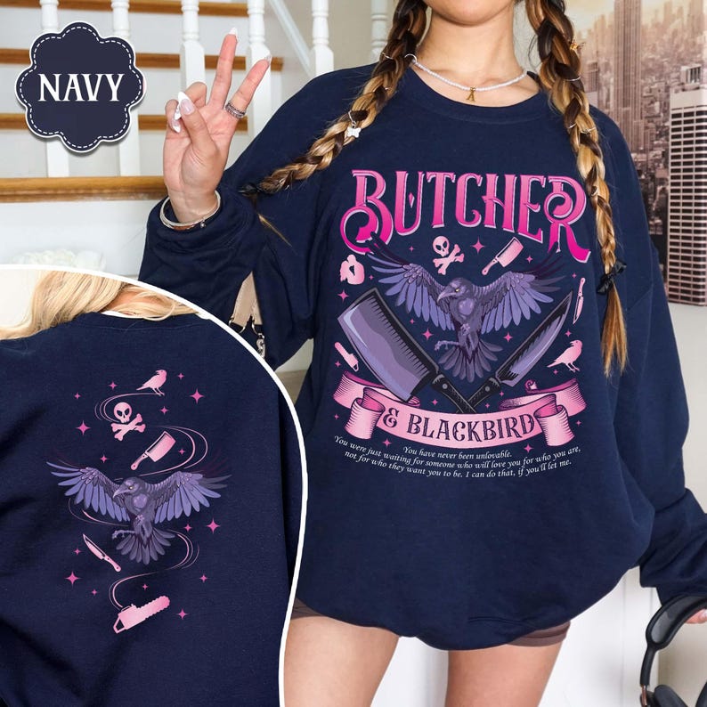 May include: Navy blue sweatshirt with a graphic design featuring the text "BUTCHER & BLACKBIRD", a raven, cleavers, and knives. The design includes pink and purple accents. The sweatshirt also has a back design with a raven, knives, and stars.