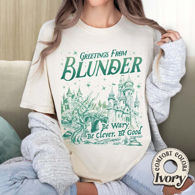 Puede incluir: Camiseta color marfil con un gr&aacute;fico verde y blanco. El gr&aacute;fico presenta las palabras "Greetings From Blunder" sobre una ilustraci&oacute;n de un castillo. Debajo, las palabras "Be Wary, Be Clever, Be Good."