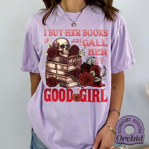 May include: A lilac-colored t-shirt with a graphic design featuring a skull atop a stack of books, surrounded by red roses. The text reads "I BUY HER BOOKS and GALL HER GOOD GIRL". The shirt has short sleeves and a relaxed fit.