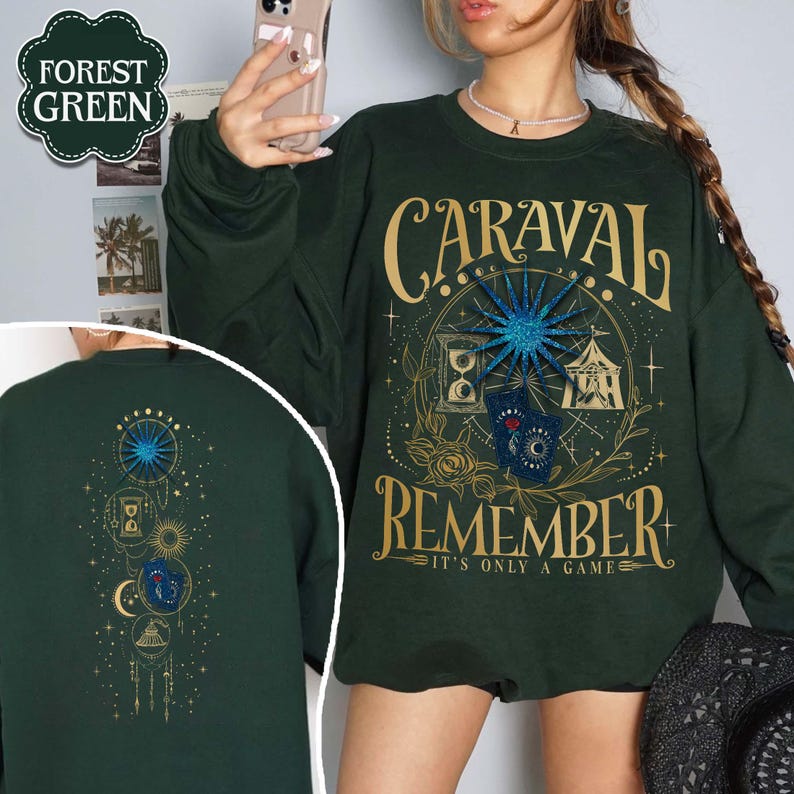 K&ouml;nnte beinhalten: Waldgr&uuml;nes Sweatshirt mit goldenem Text, der "CARAVAL REMEMBER IT'S ONLY A GAME" lautet. Das Design umfasst eine Windrose, eine Sanduhr und ein Zelt. Die R&uuml;ckseite des Sweatshirts hat ein &auml;hnliches Design mit Himmelselementen.