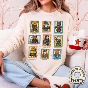 K&ouml;nnte beinhalten: Cremefarbenes Sweatshirt mit neun Katzenillustrationen, die als Briefmarken gestaltet sind. Jede Katze ist in Fantasy-Kleidung gekleidet und tr&auml;gt Namen wie "Furdo" und "Meowndalf". Das Sweatshirt hat das "Comfort Colors"-Logo.