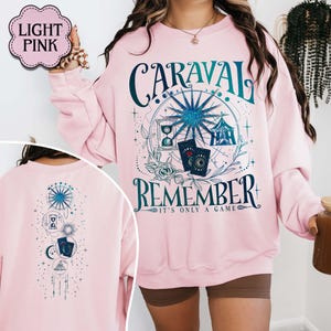 K&ouml;nnte beinhalten: Hellrosa Sweatshirt mit dem Text "CARAVAL REMEMBER" in T&uuml;rkis und Wei&szlig;. Das Design umfasst ein Riesenrad, eine Sanduhr, Spielkarten und florale Elemente. Die R&uuml;ckseite des Sweatshirts hat ein passendes Design.