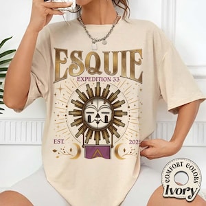 May include: Ivory-colored t-shirt with the word "ESQUIE" in a gold, vintage font. Below, the text "EXPEDITION 33" is displayed. The shirt features a sun graphic with a face, surrounded by a circular design, and the text "EST. 2025".