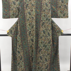 May include: A long-sleeved kimono featuring a repeating intricate paisley and floral pattern in shades of teal, dark green, rust orange, and cream. The garment is constructed with vertical panels of varying widths and is displayed on a black stand.