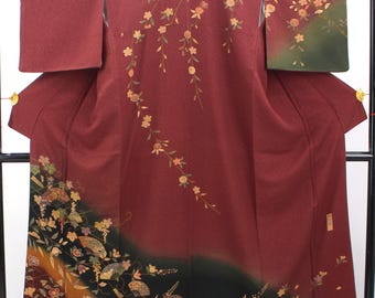 May include: A deep burgundy kimono featuring a flowing pattern of delicate gold and light-colored cherry blossom branches across the upper section, with a lower border displaying intricate fan motifs, autumn leaves, and floral arrangements.