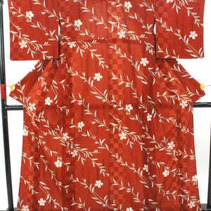 May include: A rust-red kimono featuring a subtle tonal checkered pattern throughout the fabric. The garment is decorated with a repeating diagonal motif of white five-petaled flowers and thin, light-colored leafy branches extending across the surface.