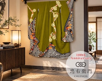 May include: An olive green kimono featuring intricate floral patterns in white, orange, and black along the sleeves and hem. The garment is displayed hanging on a black rod. A circular logo with the text 100th Anniversary and CHUEMON is visible.