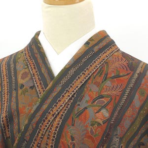 May include: Close-up of a patterned kimono with a white collar. The kimono features a mix of colors, including orange, green, and gray, with floral and striped designs. The fabric appears to be textured, and the overall style is traditional.