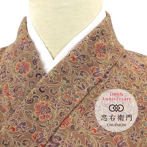 May include: A brown kimono featuring an intricate floral pattern in shades of red, purple, and green. The garment has a white inner collar and a circular logo overlay that reads 100th Anniversary, a double infinity symbol, and the text 忠右衛門 CHUEMON.