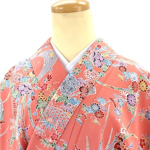 May include: A coral-colored kimono with a vibrant floral pattern. The design features detailed illustrations of flowers, leaves, and geometric shapes in shades of blue, purple, red, and green. The kimono is displayed on a mannequin.