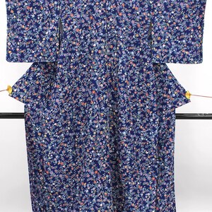 May include: A dark blue kimono featuring an all-over dense floral pattern in shades of pink, white, light blue, and orange. The garment has wide sleeves, a structured back panel, and two small gold-colored fan-shaped ornaments attached to the sides.