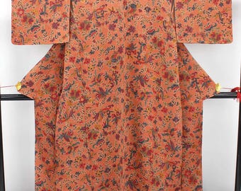 May include: A peach-colored kimono with a vibrant floral pattern. The design features red, blue, and yellow flowers and leaves. The traditional Japanese garment is displayed on a black rack, showcasing its full length and intricate details.