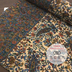 May include: A roll of fabric featuring a cream background with a dense pattern of blue, red, and green floral and paisley motifs. A circular white label is present with the text 100th Anniversary, a logo, and the Japanese characters 忠右衛門 and CHUEMON.