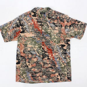 Puede incluir: Camisa de manga corta con botones y diseño estampado. La camisa presenta una mezcla de motivos florales, geométricos y ondulados en tonos negro, verde, rojo y beige. La camisa tiene un cuello clásico y un corte relajado.