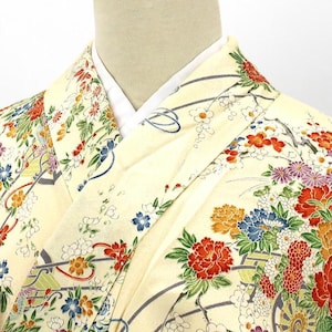 May include: Close-up of a cream-colored kimono with a vibrant floral pattern. The design features red, orange, blue, and purple flowers with green leaves. The kimono has a white collar and is likely made of silk.