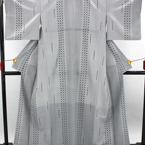 May include: A light gray kimono featuring vertical stripes of dark gray geometric patterns and scattered small, dark gray linear motifs. The garment has wide sleeves, a structured back panel, and a subtle overall textured weave throughout the fabric.