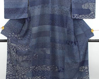 May include: A blue kimono featuring a patchwork design of various shades of blue and indigo. The garment displays horizontal bands of differing textures and patterns, including floral, geometric, and dotted motifs, with small gold accents on the sleeves.