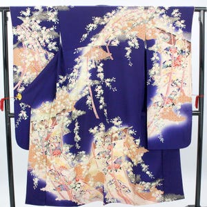 Puede incluir: Un kimono azul oscuro con un diseño floral. El kimono presenta un patrón de flores de cerezo blancas, acentos color melocotón y cintas rosas. La prenda se exhibe en un soporte negro.