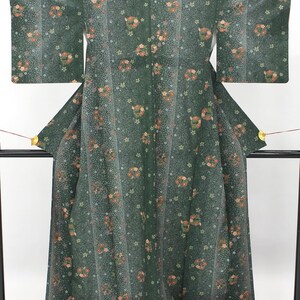 May include: A dark green kimono featuring a repeating pattern of small orange and white floral clusters, interspersed with subtle geometric lines and a fine stippled texture across the fabric, displayed on a black stand with gold accents.