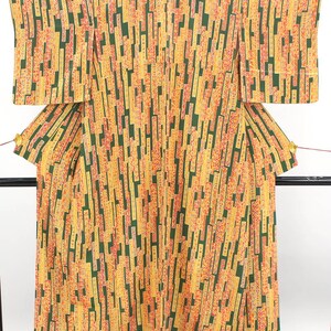 May include: A kimono featuring a repeating pattern of vertical rectangular blocks in shades of yellow, orange, and red set against a dark green background. The garment has wide sleeves and a structured shape, displayed hanging on a black frame.